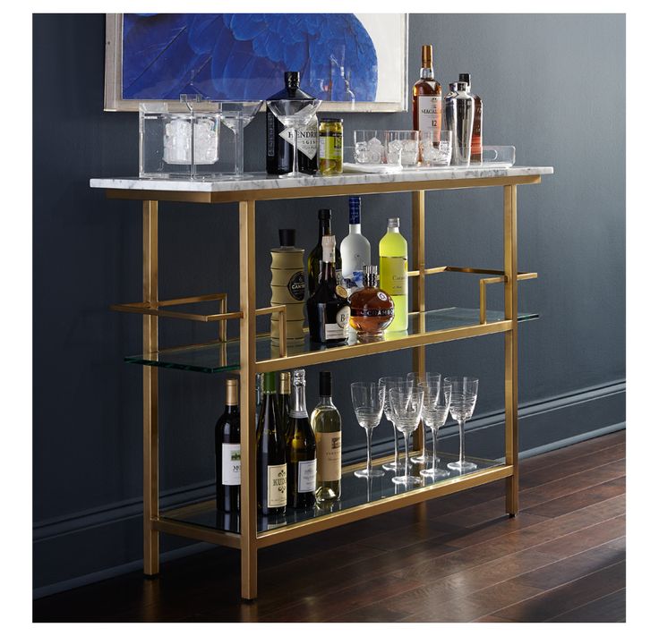 a gold bar cart with wine glasses and liquor bottles on the top, in front of a gray wall