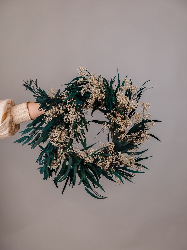 a person holding a wreath with white flowers and green leaves in their hands on a gray background