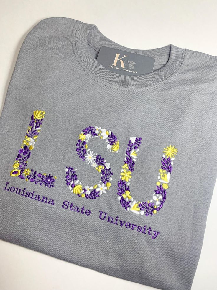 Custom University Embroidered Floral Tee. This unique shirt features delicate embroidered flowers on the letter initials of the college of your choice, adding a feminine touch to your fan gear. Gildan Spring Embroidered Gray Tops, Embroidered Short Sleeve College T-shirt, College Embroidered Short Sleeve T-shirt, Spring T-shirt With Multicolor Embroidered Text, Embroidered Short Sleeve T-shirt For College, Letter Embroidery Cotton T-shirt For College, Multicolor Embroidered Text T-shirt For Spring, Casual T-shirt With Multicolor Letter Embroidery, Casual Multicolor Embroidered Letter T-shirt