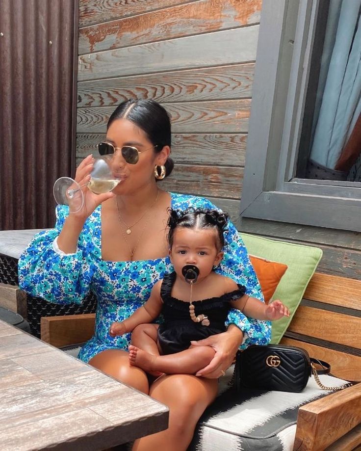 a woman sitting on a bench drinking from a wine glass with a baby in her lap