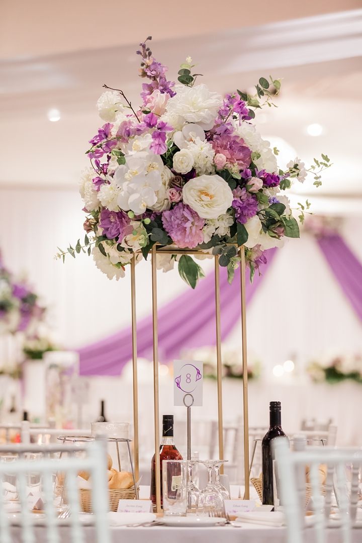 an elegant centerpiece with purple and white flowers is displayed on a table at a wedding reception