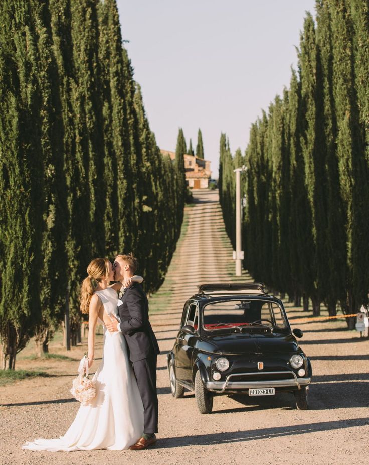 a bride and groom kissing in front of an old car on the side of a road