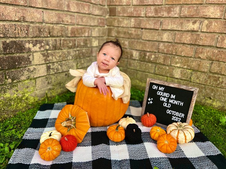 a baby in a pumpkin costume sitting on a checkered blanket with other pumpkins and gourds