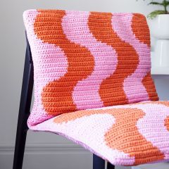 an orange and pink crocheted pillow sitting on top of a chair next to a potted plant