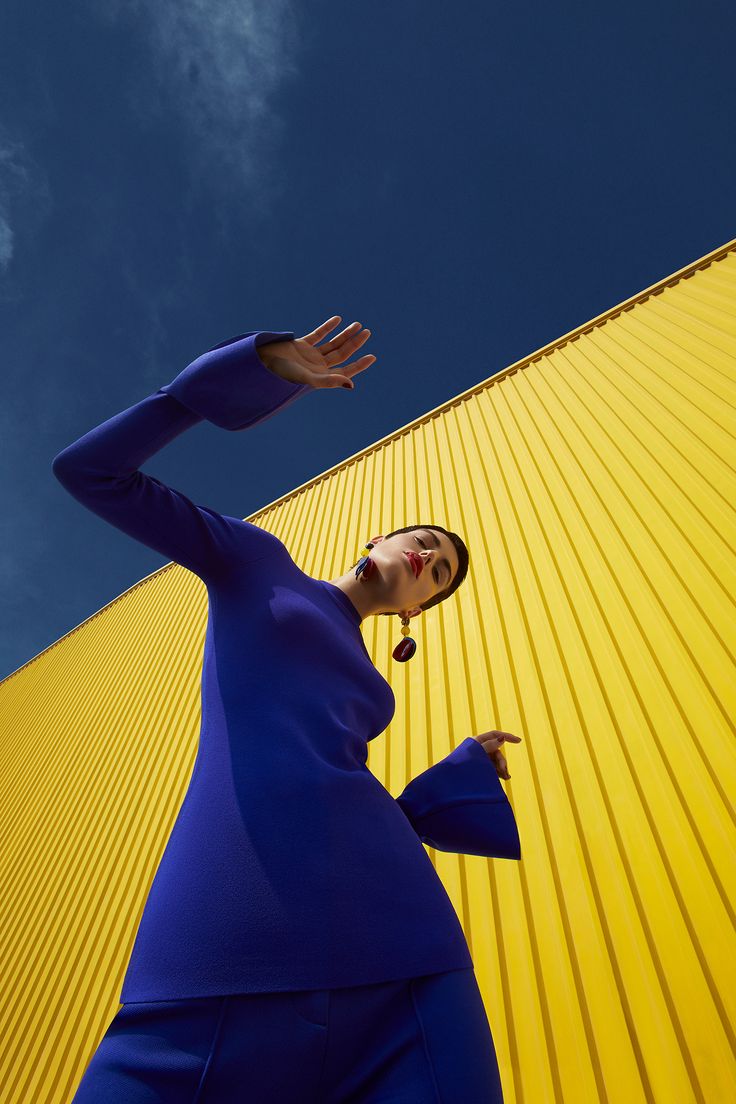 a woman in a blue dress standing next to a yellow building with her hand up