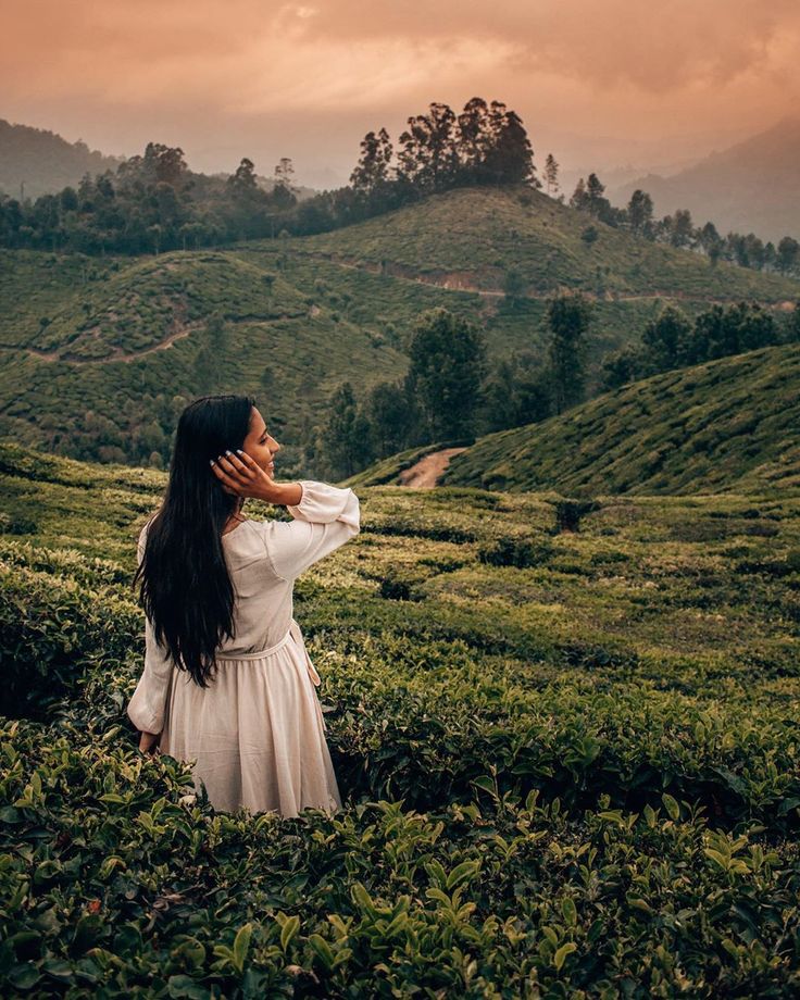 a woman standing in the middle of a lush green field with mountains behind her and clouds overhead