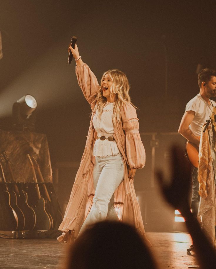 a woman standing on top of a stage holding her arms up in the air while two other people stand behind her