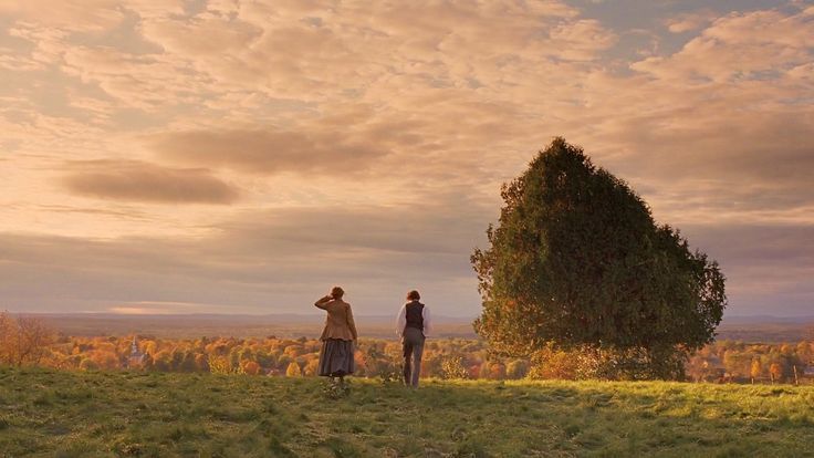 two people standing on top of a grass covered hill next to a tree with clouds in the background