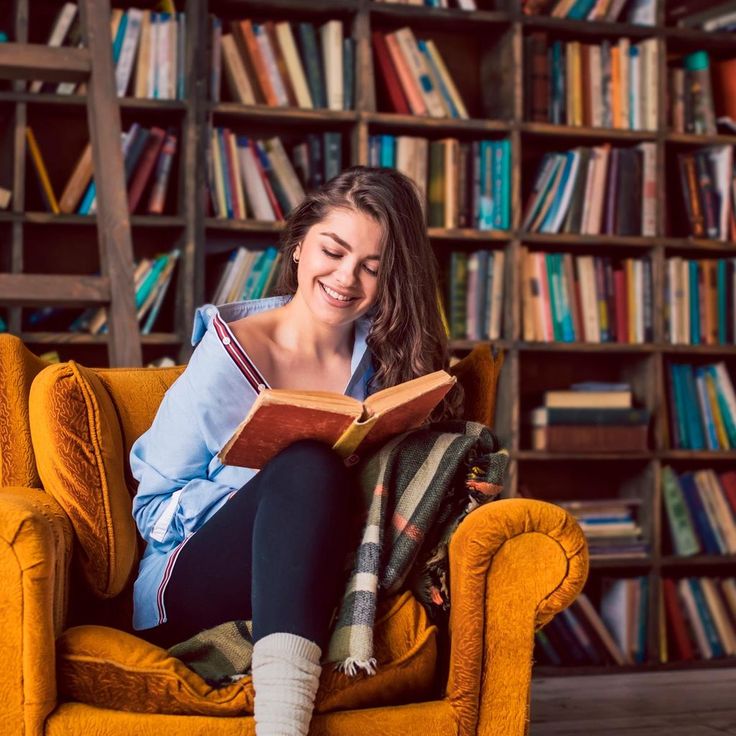 a woman sitting on a chair reading a book in front of a bookshelf