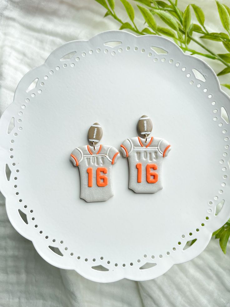 two orange and white football jersey earrings sitting on top of a white plate next to green leaves