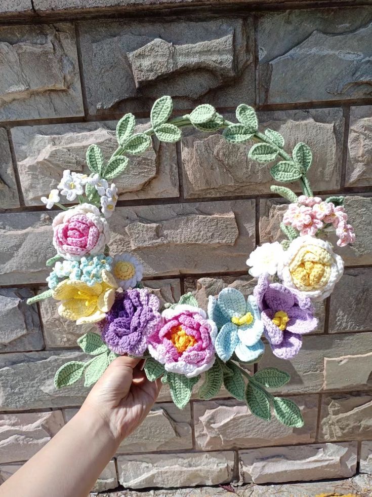 crocheted wreath with flowers and leaves in front of a brick wall, being held up by a hand