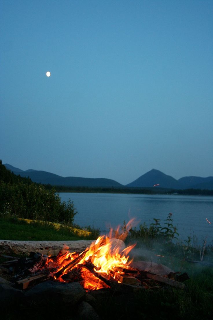 a campfire with the moon in the sky above it and water behind it at night