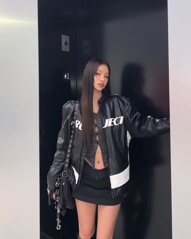 a woman standing in front of a black wall with her hand on her hip, wearing a leather jacket and shorts