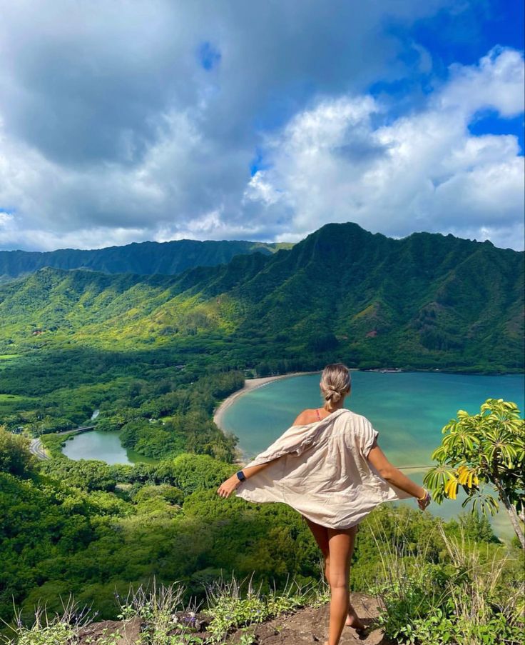 a woman in a dress is walking up a hill towards the water and hills behind her