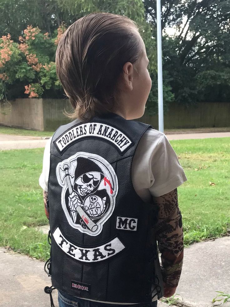 a young boy wearing a leather vest with patches and tattoos on his arm, standing in front of a grassy area