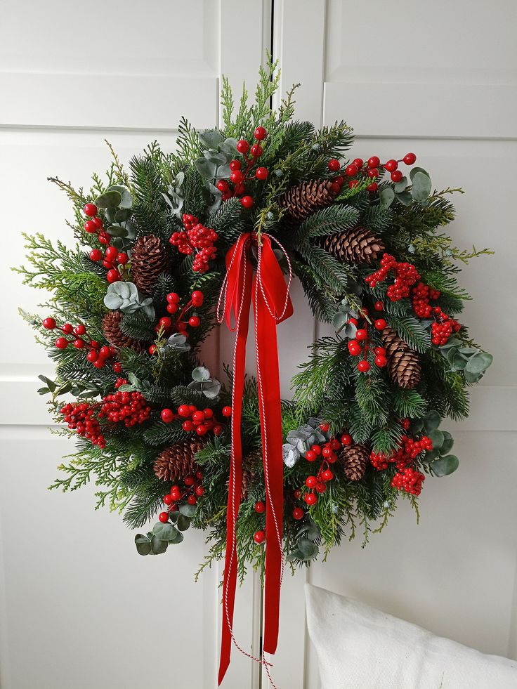 a christmas wreath with red berries and pine cones hanging on a white door front porch