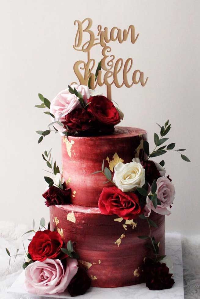 a three tiered cake with red and pink flowers on top, topped with a wooden name sign