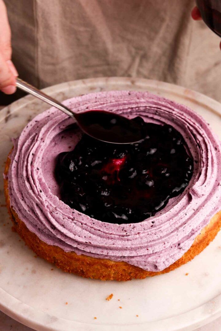 a person holding a spoon over a cake with purple frosting on it and blueberries in the middle