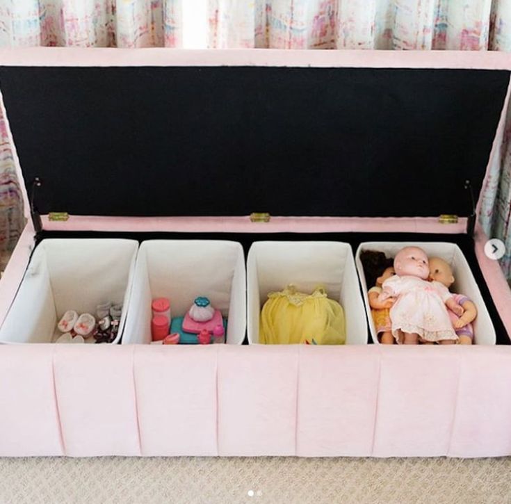 an open pink suitcase filled with dolls and other toys on a carpeted floor next to curtains