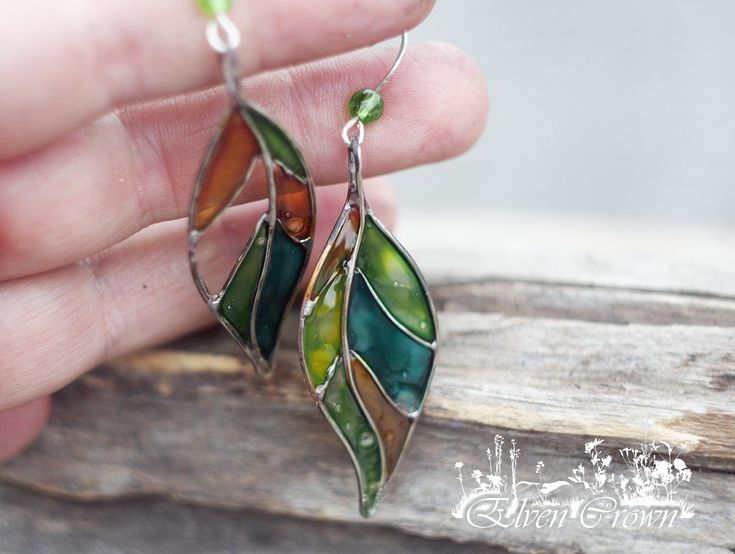 a pair of green, orange and brown leaves hanging from silver earwires on a piece of wood