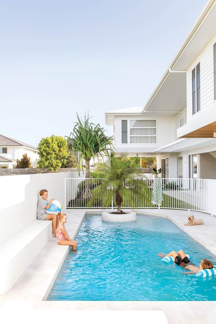 two people sitting on the edge of a swimming pool in front of a white house