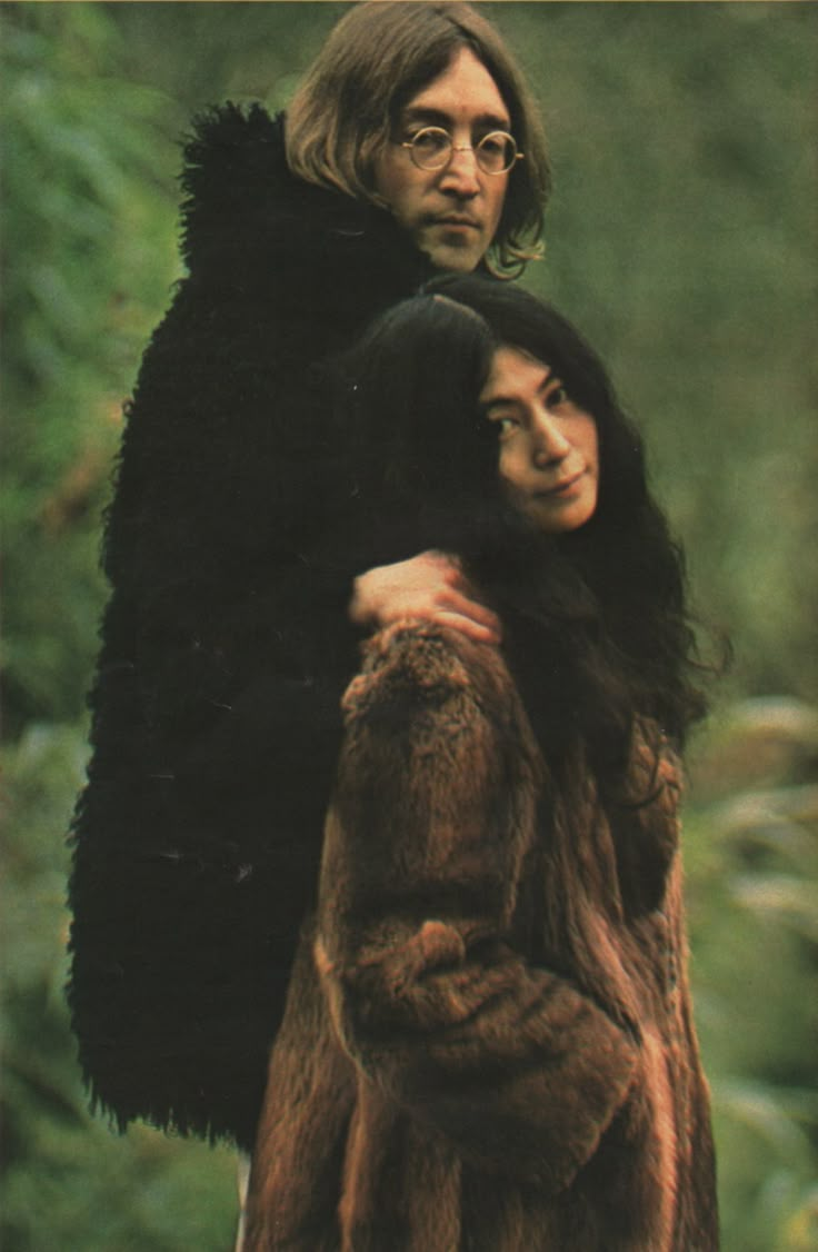 two people standing next to each other in front of trees and bushes, one woman is wearing a fur coat