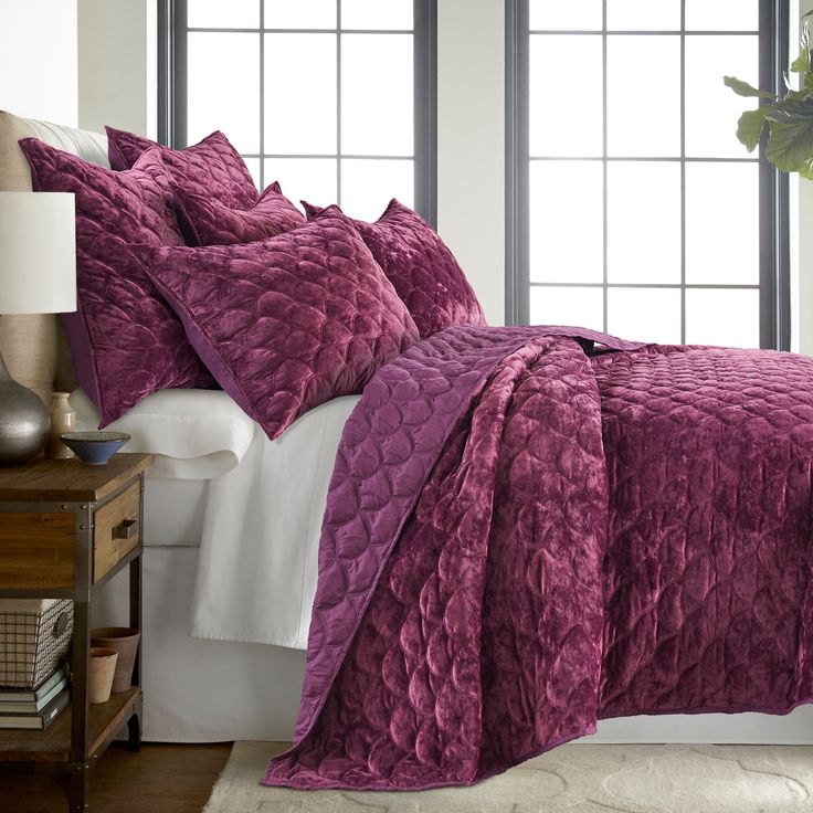 a bed with purple comforter and pillows in a room next to a window,