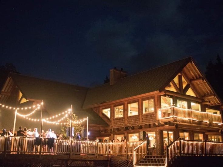 people are standing on the porch of a large log cabin at night with christmas lights