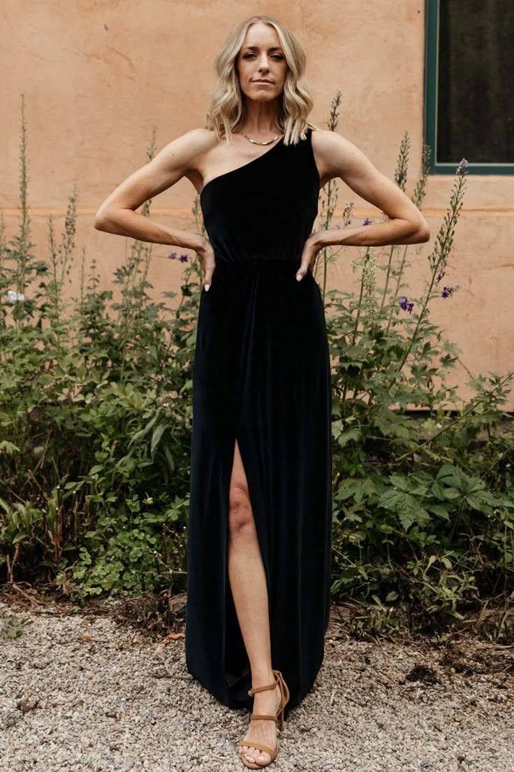 a woman standing in front of a building wearing a black one shoulder dress and sandals