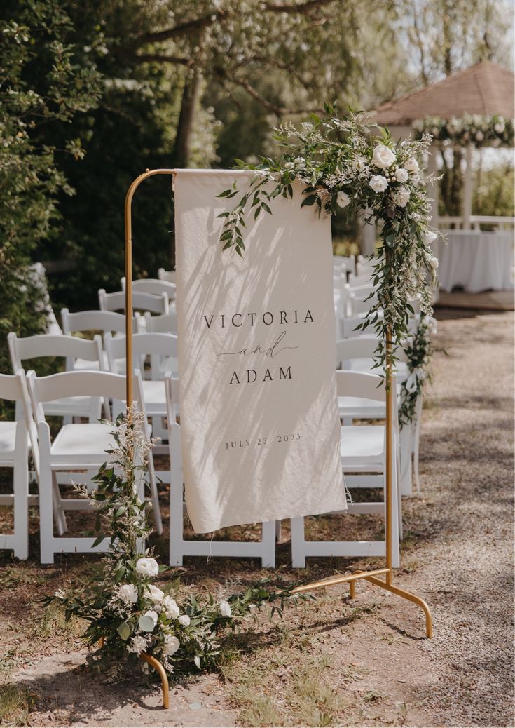 an outdoor wedding ceremony with white chairs and greenery on the aisle, sign that says victoria and adam