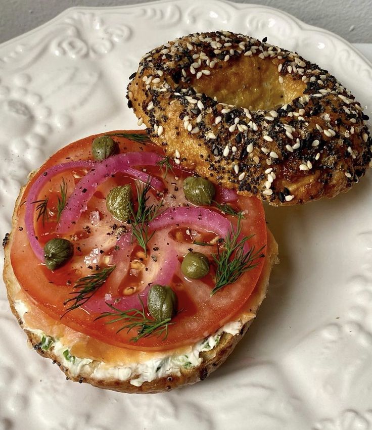 a bagel sandwich with tomatoes, onions and sprouts on it sitting on a white plate