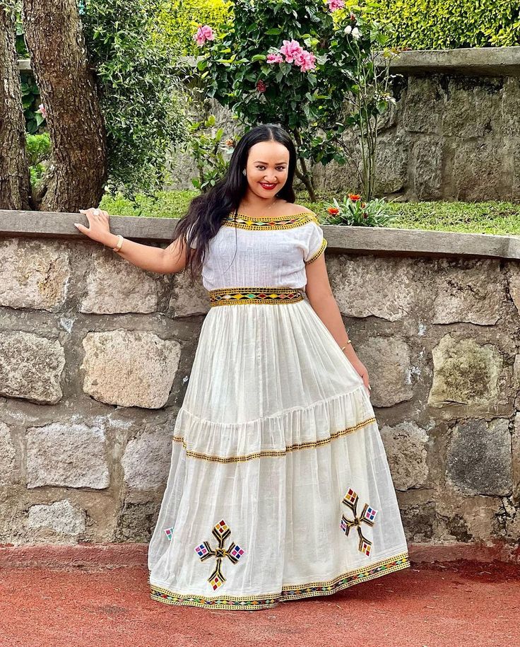 a woman standing next to a stone wall wearing a white dress and holding her arms out