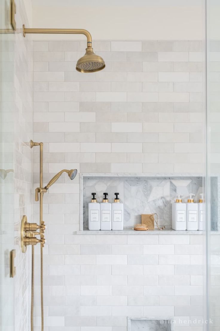 a white tiled bathroom with gold fixtures and marble counter tops, shower head, soap dispenser, and shelves