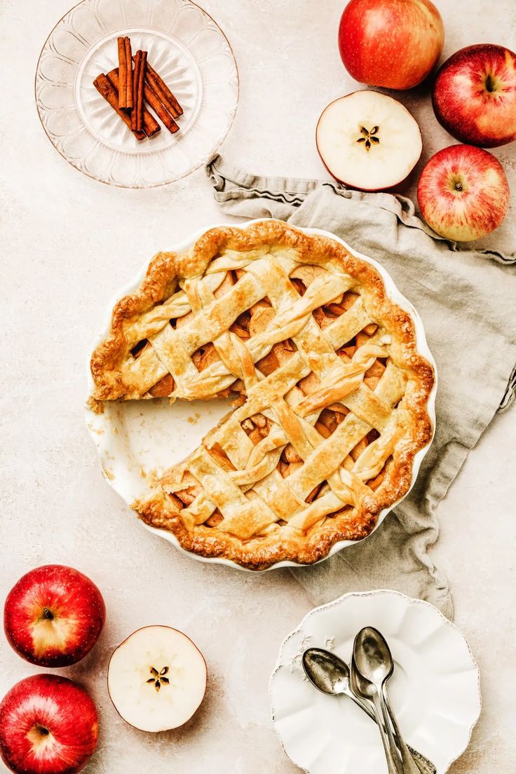 an apple pie on a table with apples and cinnamon sticks next to the pie plate