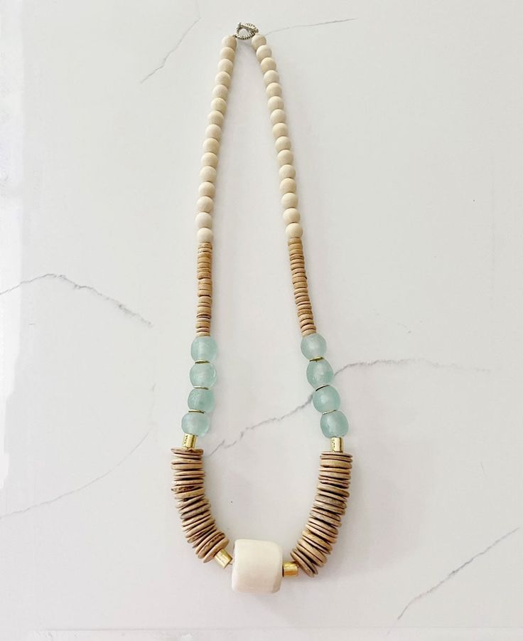 This eye-catching necklace beautifully blends earthy coconut wood, and recycled glass beads from Ghana, creating a piece that exudes natural elegance and artisanal craftsmanship. Design Feature: Crafted with a mix of tan coconut wood disk beads, white round wood beads, clear aqua recycled glass beads, and a large white barrel bead. Material: Made with high-quality materials including wood, and recycled glass, strung on an antiqued bronze hammered toggle closure. Size: Measures 32.5 inches in tot Long Wood Bead Necklace, Earthy Necklace, Beach Jewellery, Coconut Wood, Tassels Decor, Aqua Glass, Recycled Glass Bead, Wood Bead Necklace, Tin Gifts