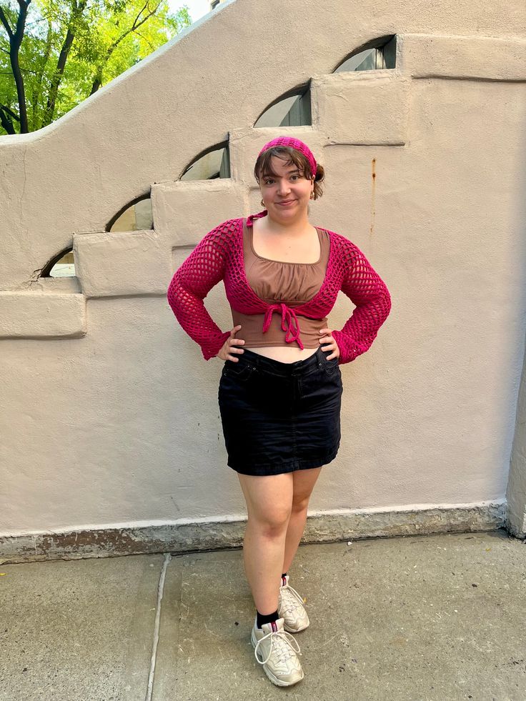 a woman standing in front of a white wall wearing a pink top and black skirt