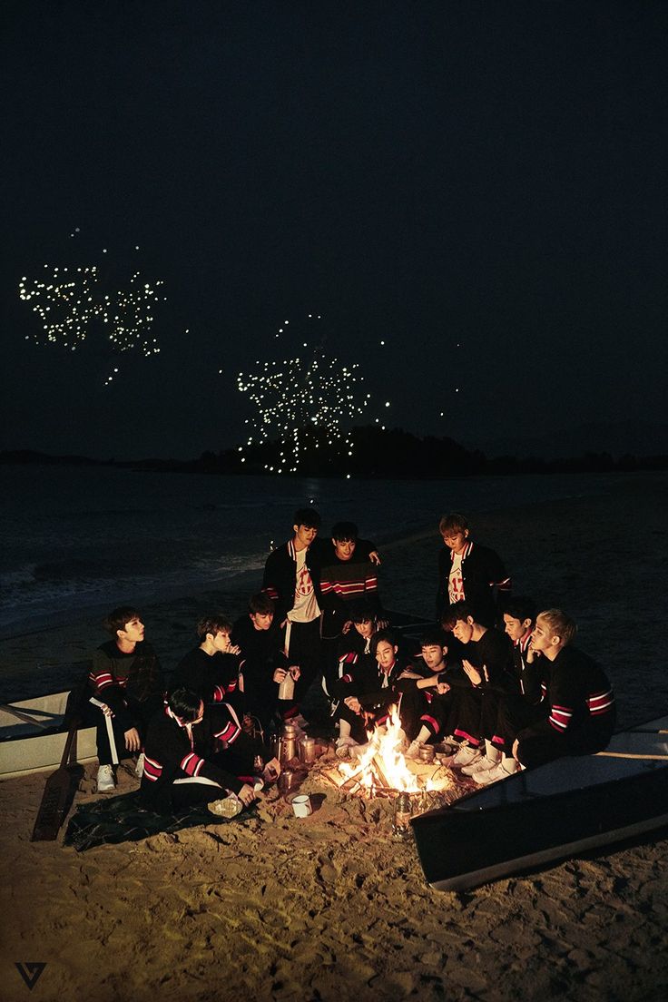 a group of people sitting around a campfire on the beach with fireworks in the sky