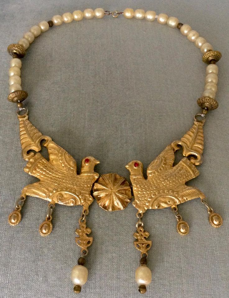 You are looking at an absolutely exquisite IMPORTANT VINTAGE CADORO ETRUSCAN BYZANTINE REVIVAL INSPIRED RICH GILT GOLD-TONE METAL MASSIVE 7" LONG BIB PENDANT COLLAR CHOKER NECKLACE SHOWCASING A STUNNING DIMENSIONAL SCULPTURAL CENTERPIECE IN THE SHAPE OF A PAIR OF GORGEOUS AN GIANT LOVEBIRDS WITH VIBRANT RUBY RED GLASS CABOCHON EYES FACING EACH OTHER, FEATURING A CHAIN CONSISTENT OF GLASS BAROQUE FAUX PEARLS AND GOLD-TONE METAL MELLON BEADS, EXECUTED WITH EXTREME DETAIL, UNSIGNED, Circa 1980's. AN ABSOLUTELY ICONIC PIECE OF JEWELRY AND A TRUE SHOWSTOPPER EXTRAORDINARILY BEAUTIFUL AND ELEGANT, EXTREMELY RARE AND ALMOST IMPOSSIBLE TO FIND THIS BEAUTIFUL COLLAR CHOKER BIB HINGED PIECE FEATURES A DELICATELY SCULPTED GIANT COUPLE OF GOLD METAL LOVEBIRDS WITH RUBY RED GLASS EYES AND DANGLING CHAR Designer Runway, Collar Choker, Bib Necklaces, Choker Collar, Pearl Charms, Vintage Designer, Red Glass, Baroque Pearls, Ruby Red