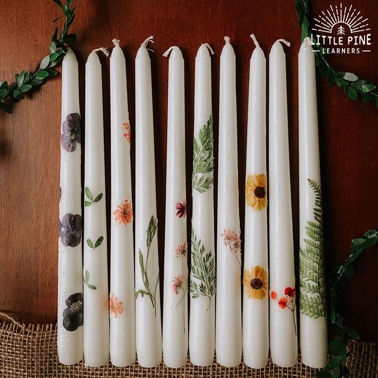 six white candles with floral designs on them sitting on a table next to greenery