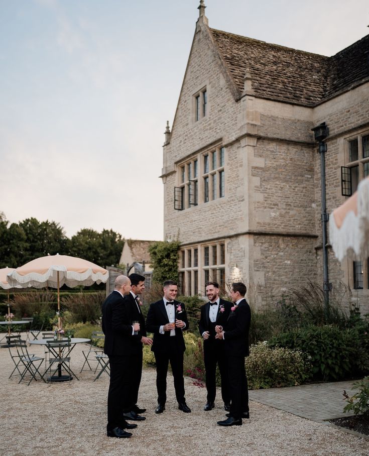 three men in tuxedos are standing outside an old brick building with tables and umbrellas