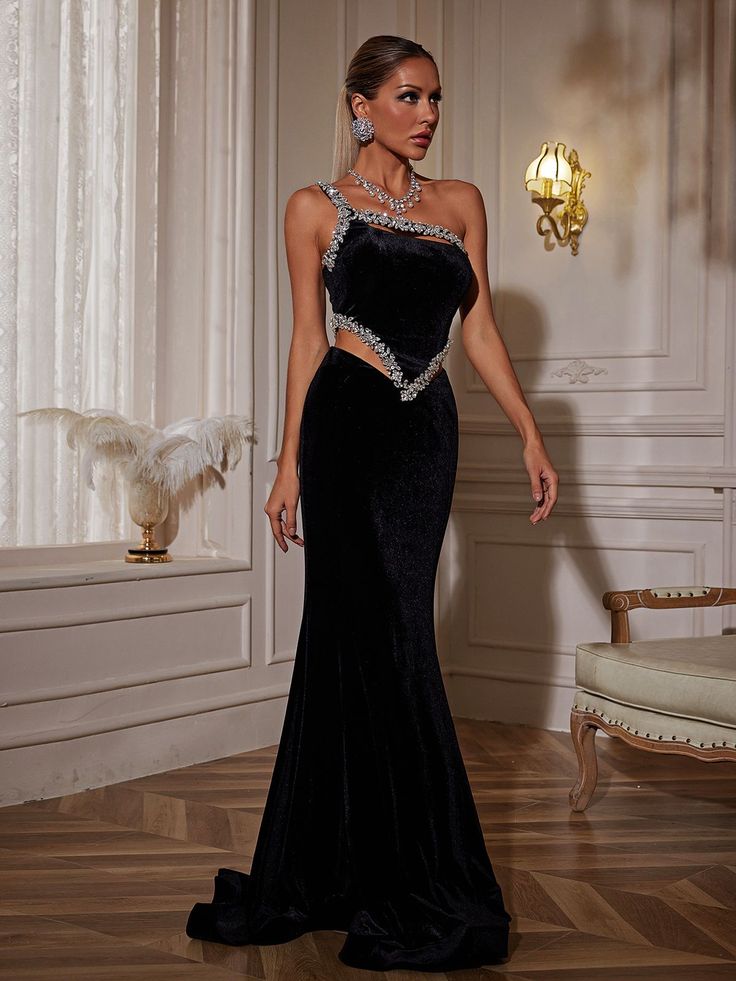 a woman in a black evening gown standing on a wooden floor with a chandelier behind her