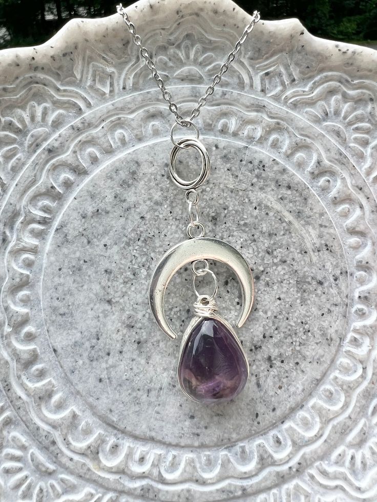 This statement necklace features a gorgeous large amethyst gemstone. The amethyst is wire wrapped by hand with silver wire and bezel set on a silver pear shaped base. The stone hangs from a brilliant silver moon adding a celestial element to the pendant. The moon is suspended from a dainty antique silver twist hoop. The deep purple stone is domed on top and flat on the back. Many dark and light shades of purple are displayed throughout the stone. The color variations were created by the minerals that grew with the amethyst in nature. The stunning pendant is 63.3 mm long and is attached to a beautiful silver stainless steel chain with a lobster clasp. Amethyst is a grounding, protective, calm stone that encourages balanced energy, love of the divine, and selflessness. * Information for meta Spiritual Crescent Wire Wrapped Necklaces, Silver Crescent Necklace With Natural Stones, Mystical Sterling Silver Crystal Necklaces With Natural Stones, Mystical Sterling Silver Necklace With Natural Stones, Purple Amethyst Wire Wrapped Crystal Necklace, Purple Amethyst Wire Wrapped Necklace, Silver Crescent Necklace For Healing, Purple Amethyst Wire Wrapped Necklaces, Bohemian Wire Wrapped Amethyst Necklace