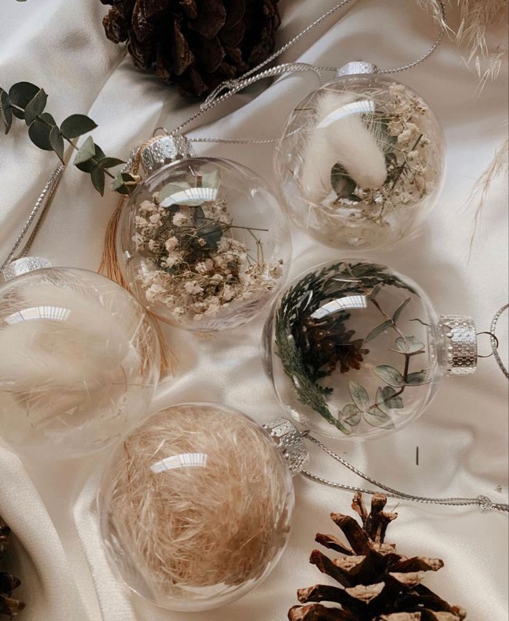 four glass ornaments with pine cones and greenery in them on a white table cloth