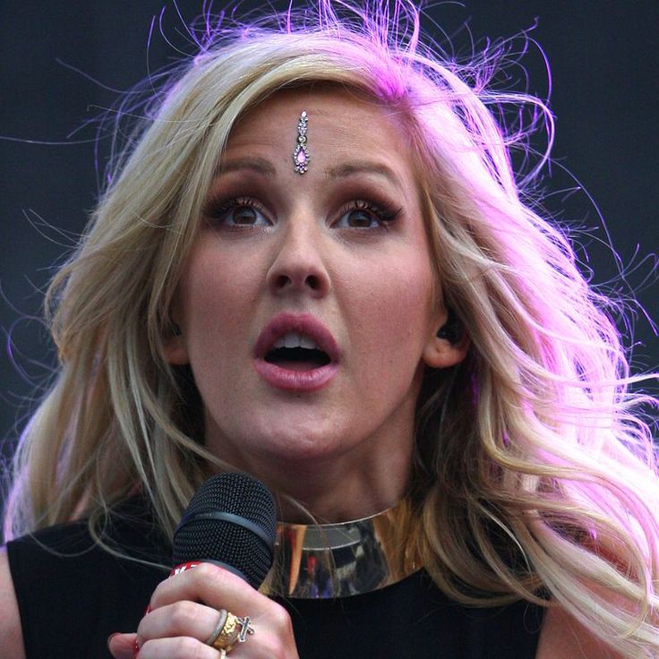 a woman with blonde hair holding a microphone in her right hand and wearing a choker around her neck