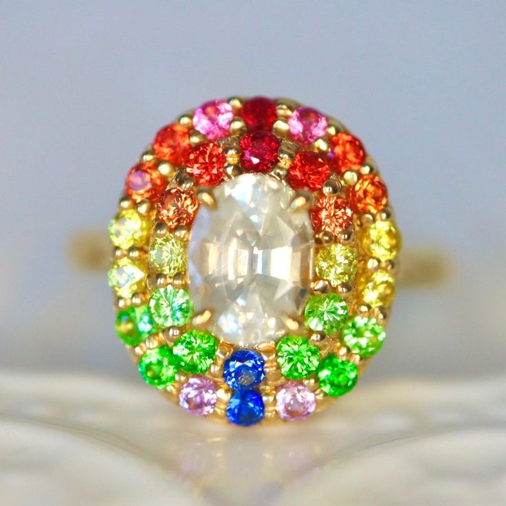 Opalescent White Sapphire and rainbow color halo ring Condition: Brand New & Custom Designed by me Size: US 7 sizable Metal: Solid 14K Yellow Gold Center Stone: Natural White Sapphire. Opalescent white color. Internally flawless.  Side Stones: Red rubies, orange sapphires, neon orange sapphires, yellow sapphires, mint garnets, tsavorite garnet, blue sapphires, purple sapphires 1.5ct in total. Measurement: 15*13mm face. 7mm height. Band Width: 2.4mm on the back Band Thickness: 1.4mm on the back C Multicolor Jewelry As A Gift, Multicolor Oval Heirloom Ring, Multicolor Halo Ring As Gift, Multicolor Halo Rings As A Gift, Multicolor Halo Rings For Gift, Wedding Rainbow Multi-stone Rings, Multicolor Halo Jewelry For Wedding, Heirloom Multicolor Oval Rings, Heirloom Style Multicolor Oval Rings