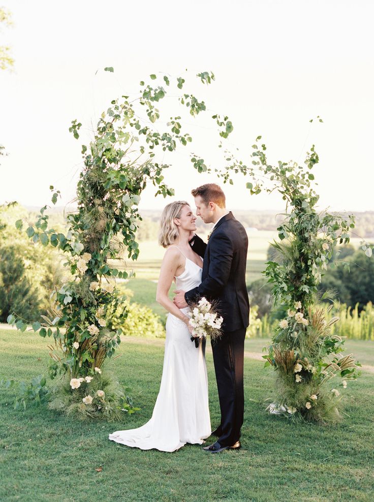 a bride and groom standing under an arch with greenery at their wedding ceremony in the countryside