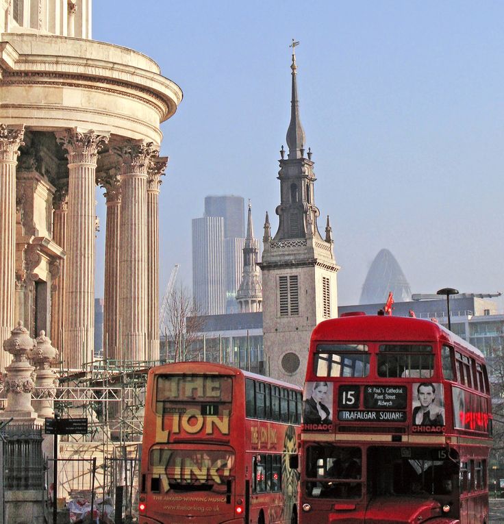 two red double decker buses driving down the street in front of an old building with spires