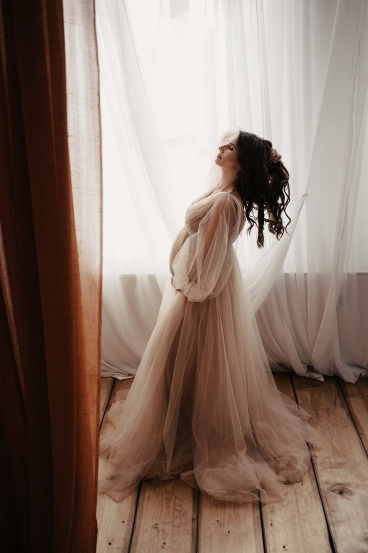 a woman standing in front of a window wearing a sheer white dress with long sleeves