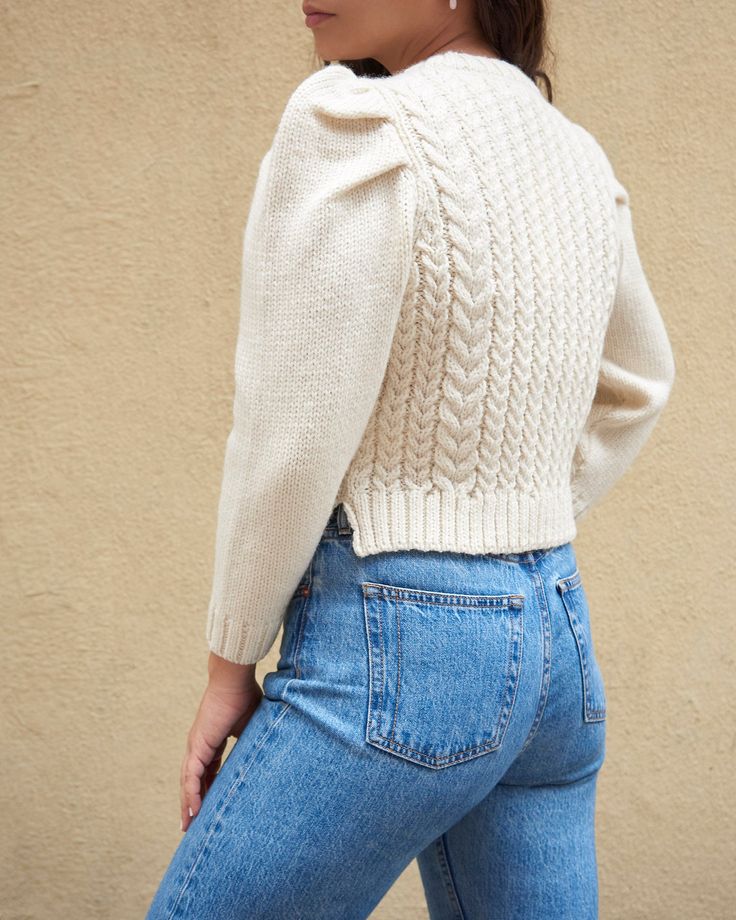 Puff-sleeved crewneck cable knit sweater in cream-colored wool. Features a slightly cropped fit and 3/4 length sleeves. Size Body Length Across Shoulders XS 16.75" 15.5" S 17.75" 16.5" M 18.75" 17.5" L 19.75" 18.5" Fitted Turtleneck Cardigan With Textured Knit, Beige Knit Turtleneck Cardigan, Beige Cable Knit Turtleneck Cardigan, Beige Cotton Cable Knit Outerwear, Cream Wool Sweater For Fall, Classic Beige Chunky Knit Outerwear, White Merino Wool Cardigan For Fall, Fitted Chunky Knit Wool Outerwear, Fitted Merino Wool Cable Knit Outerwear