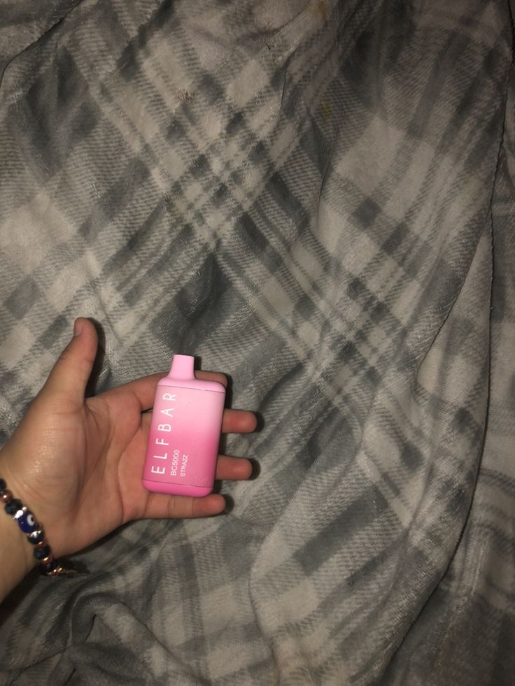 a hand holding a pink bottle on top of a bed next to a plaid blanket